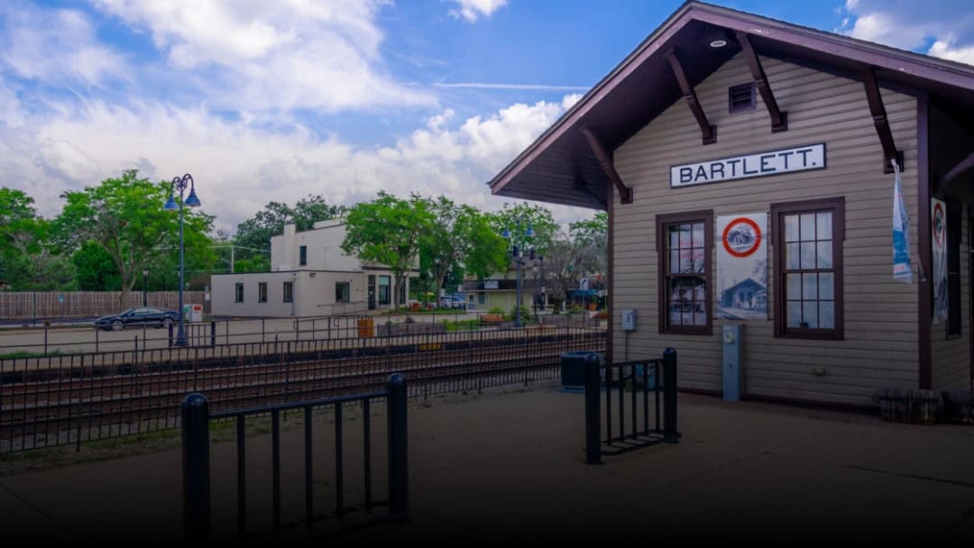 Historic Bartlett train station building set against a backdrop of train tracks, trees, and buildings